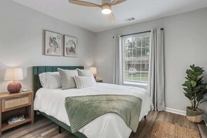 Une chambre à coucher comprenant un lit, des tables de chevet, un ventilateur de plafond, une fenêtre avec des rideaux et une plante en pot.