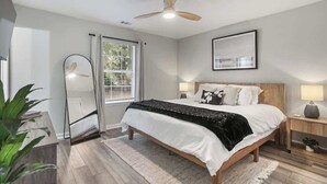 A bedroom with a bed, a ceiling fan, a window with curtains, a bedside table with a lamp, and a mirror.