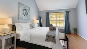 Une chambre à coucher comprenant un lit, des tables de chevet, un ventilateur de plafond, une fenêtre avec des rideaux et une œuvre d’art accrochée au mur.