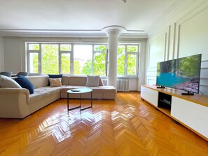 A modern living room with a sectional sofa, a coffee table, and a flat-screen TV on a wooden TV stand. Large windows provide natural light and a view of greenery outside. - Time Traveller's Mansion in Fatih (Istanbul)