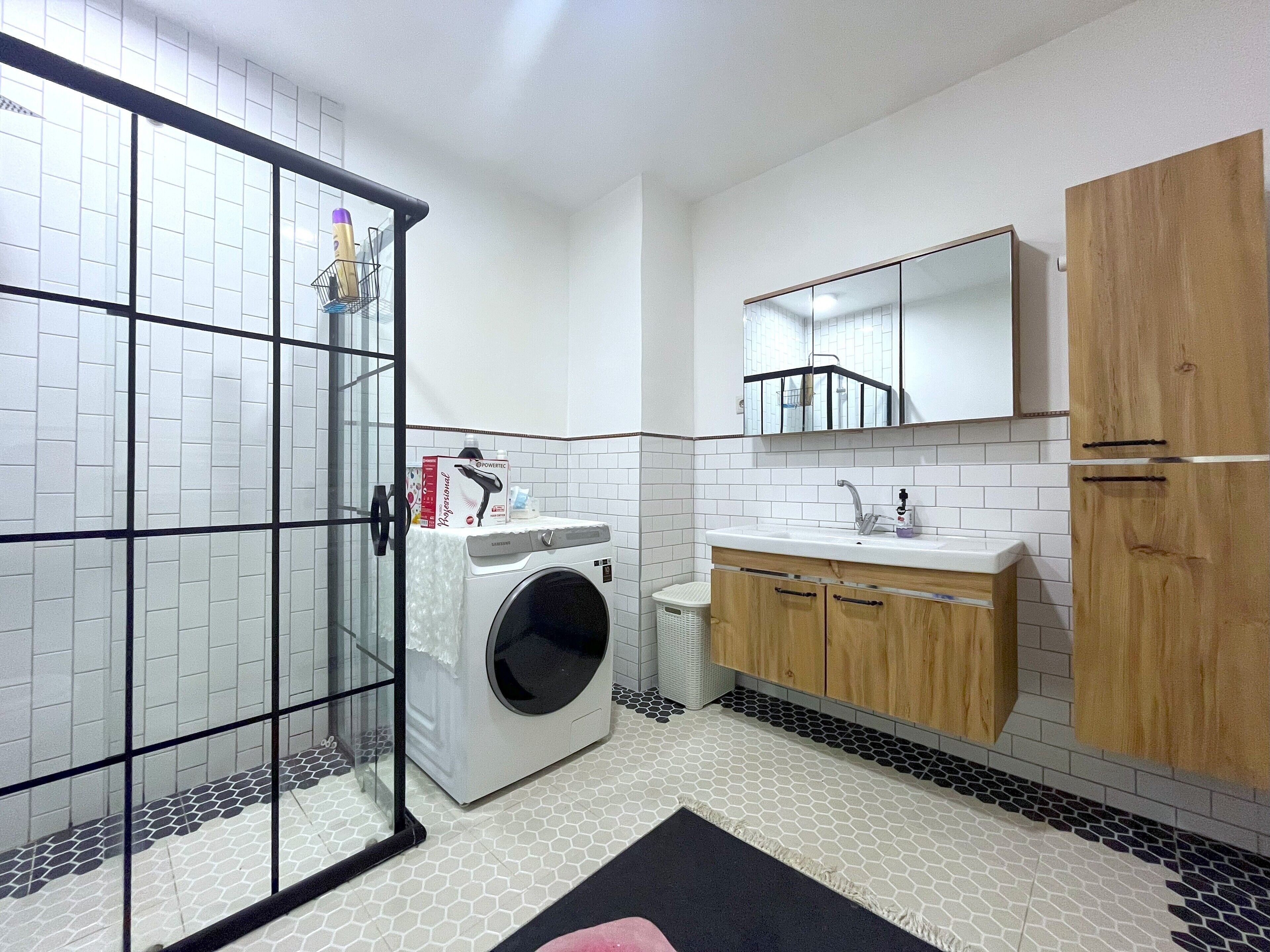 A compact bathroom with a glass shower enclosure, a washing machine, a sink with a mirror, and wooden cabinets.