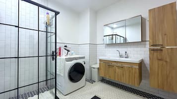 A compact bathroom with a glass shower enclosure, a washing machine, a sink with a mirror, and wooden cabinets.