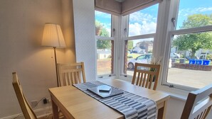 A dining area with a wooden table, four chairs, a lamp, and large windows with a view of the outside. - Bright 1 bedroom Apartment in Drymen (Drymen)