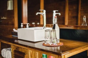 Ein Badezimmer-Waschtisch aus Holz mit einer weißen Spüle, zwei Wasserhähnen und einer Shampoo-Flasche.