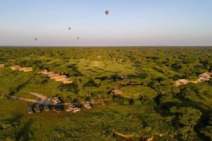 Vista aérea de um lodge de safári com várias tendas e veículos, cercado por vegetação densa e balões de ar quente ao longe.