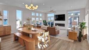 A modern living room with a dining area, large windows, a fireplace, and a ceiling fan.