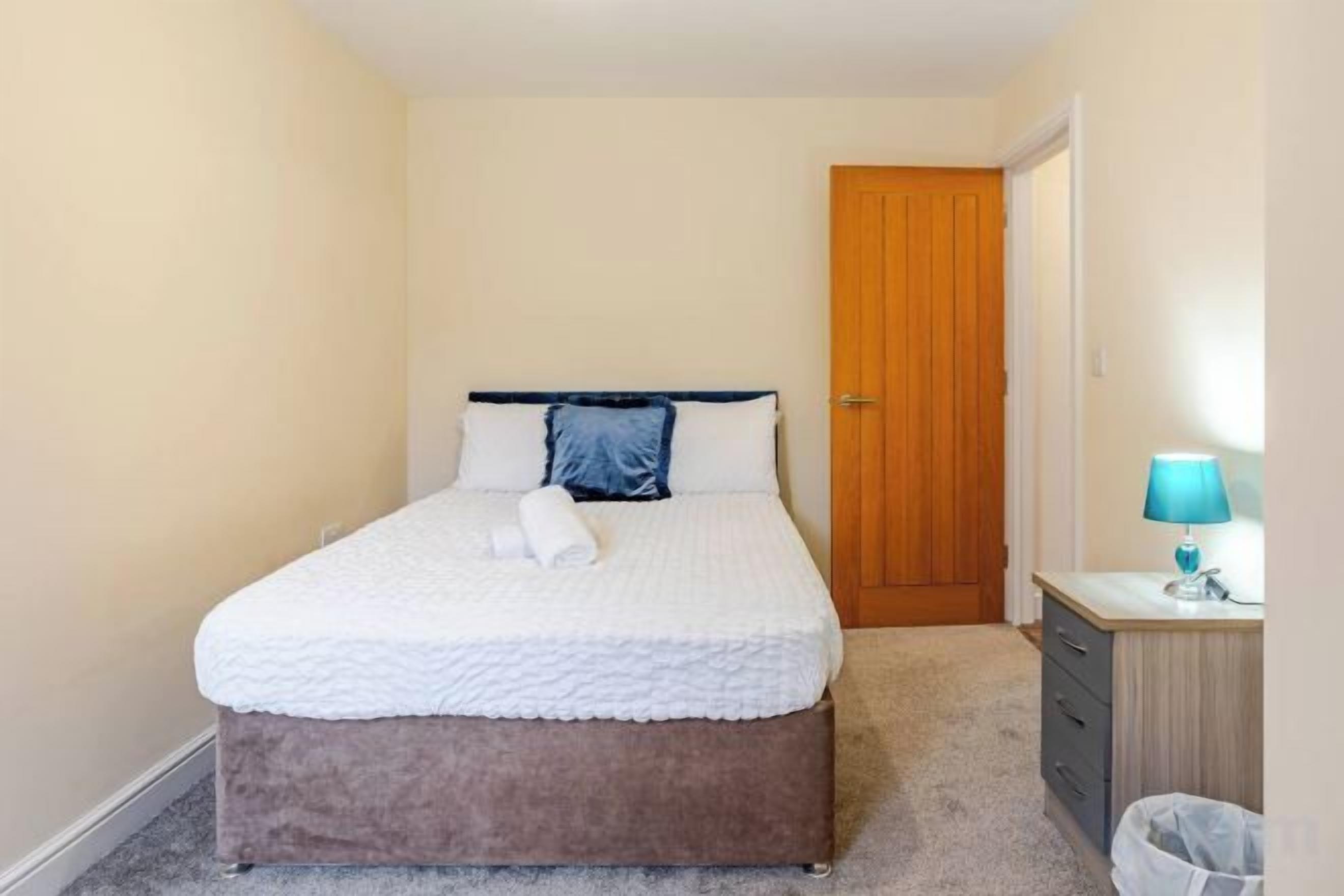 A bedroom with a bed, a wooden door, a bedside table with a lamp, and a trash bin.