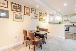 A dining area with a table and chairs, a kitchen in the background, and framed pictures on the wall. - Cosy Modern Flat, Cockermouth, Sleeps 4 (Cockermouth)