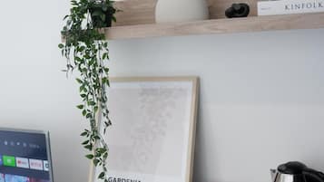 A room with a framed picture, a plant, a shelf with books, and a desk with snacks and a kettle.