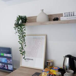 Ein Zimmer mit einem gerahmten Bild, einer Pflanze, einem Bücherregal und einem Schreibtisch mit Snacks und einem Wasserkocher.