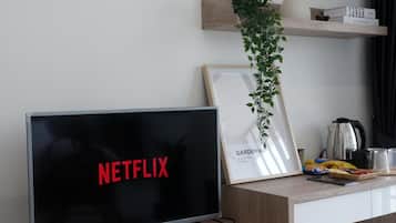 A modern living room with a flat-screen TV displaying 'NETFLIX' on the screen, a framed picture with 'GARDENIA' text, a potted plant, and a coffee maker on the counter.