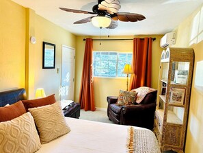 A bedroom with a bed, a chair, a ceiling fan, a window with curtains, and a wall-mounted mirror.