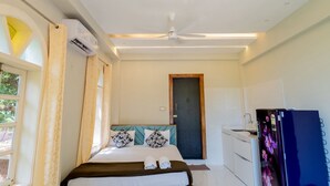 Quarto de hotel com uma cama, um ventilador de teto, janela com cortinas, uma porta e uma geladeira.