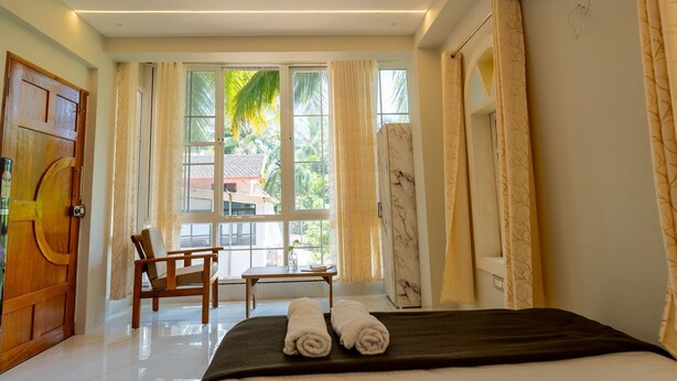 Quarto de hotel com cama, uma cadeira, janela com vista para palmeiras e porta com puxador de madeira.