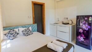 Quarto de hotel com cama, porta de madeira, cozinha compacta e geladeira com design floral.