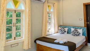 Quarto com cama de madeira, duas janelas com cortinas, aparelho de ar condicionado e ventoinha no teto.