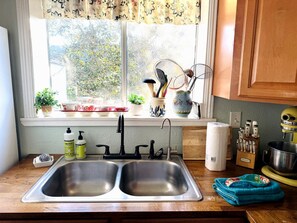 A kitchen with a double sink, a window with plants, and various kitchen utensils on the windowsill. - Pet Friendly Breathtaking Cottage with Great Views and Hot-tub in Rainbow, California (Fallbrook)