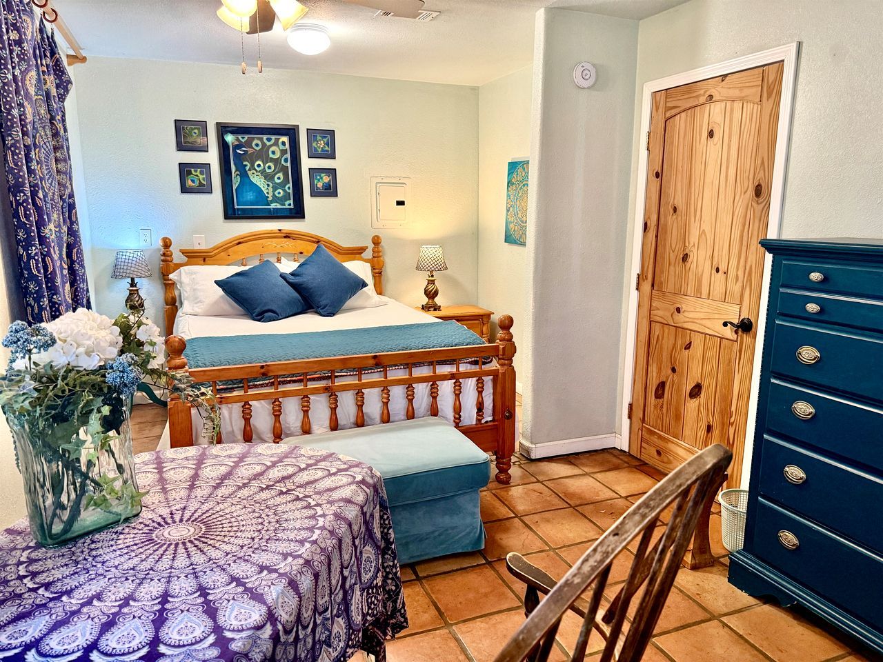 A bedroom with a double bed, a wooden wardrobe, a chair, a bedside table with a lamp, and a vase with flowers.
