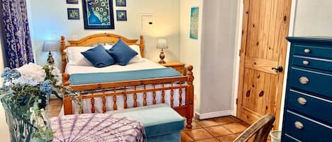 A bedroom with a double bed, a wooden wardrobe, a chair, a bedside table with a lamp, and a vase with flowers.