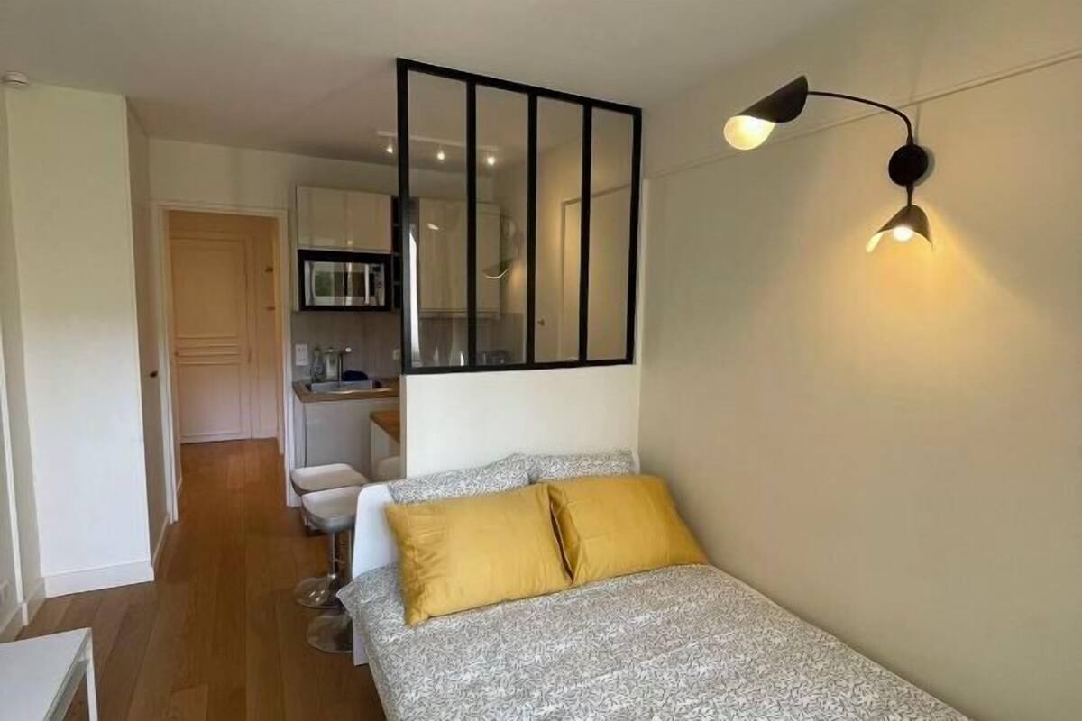 A bedroom with a bed, a bedside lamp, a mirror, and a view into the kitchen.