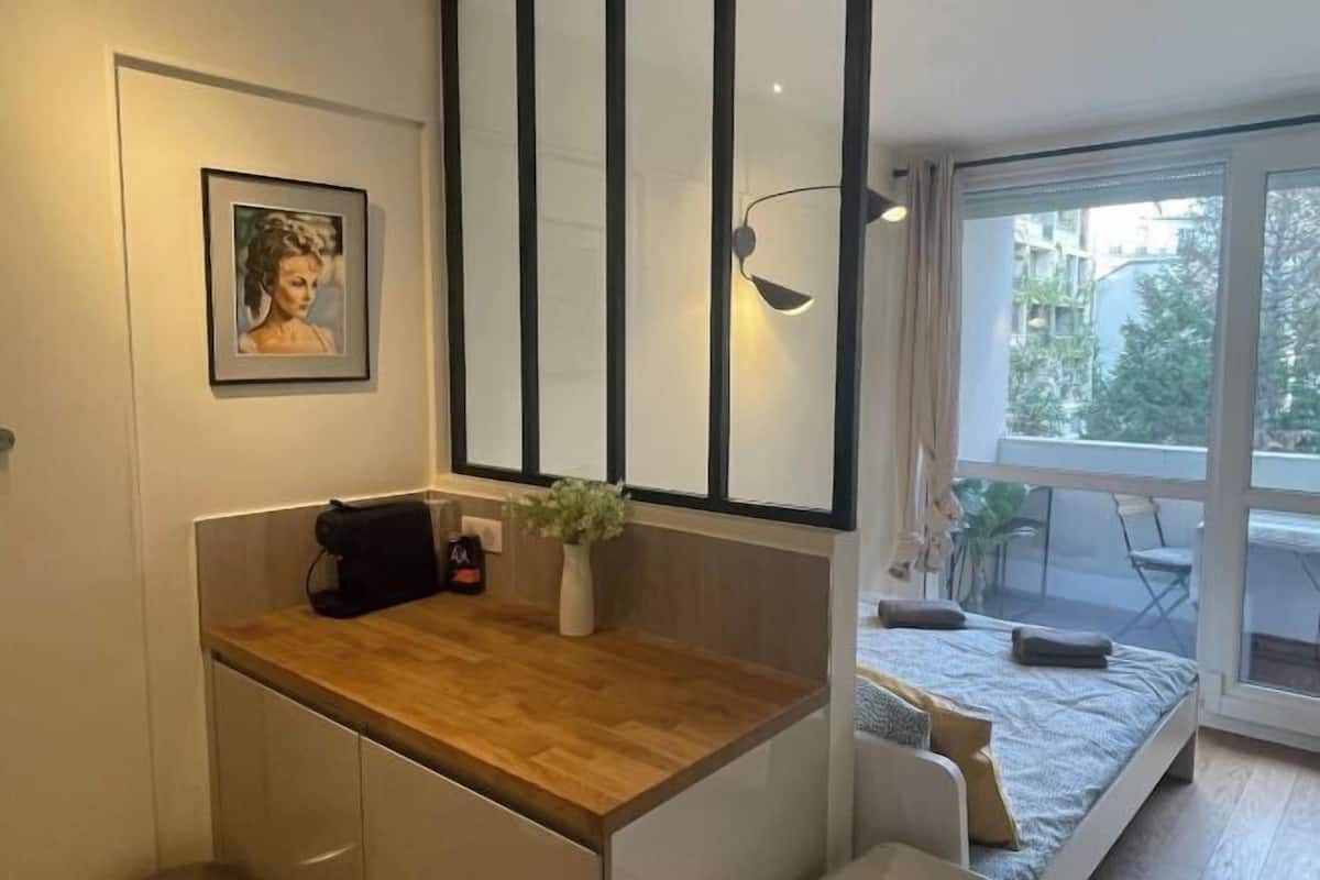 A modern bedroom with a bed, a small desk, a chair, a window with a view of the outside, and a wall-mounted light fixture.