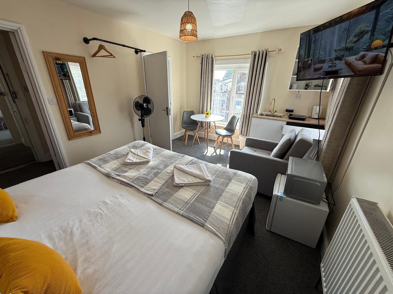 Habitación de hotel con cama, sofá, televisor, ventilador, pequeña cocina americana y ventana con cortinas.