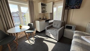 A compact living space with a kitchenette, a dining area with a table and chairs, a sofa, a TV mounted on the wall, and large windows with curtains. - Studio 32-Free Private Parking (Portsmouth)