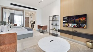 A modern hotel room with a large bed, a dining area, a TV wall, and a small kitchen.