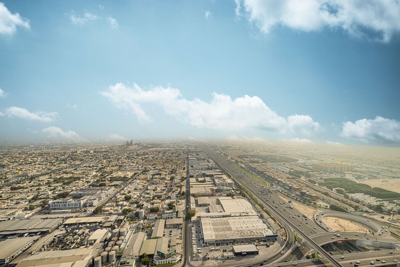A wide aerial view of a cityscape with a prominent highway, industrial buildings, and a large open area with sparse vegetation.