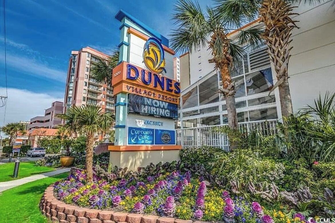 Un panneau indiquant le « Dunes Village Resort », avec une affiche signalant des chambres disponibles, un café-bar appelé « Captun's Cafe & Bar », et un jardin aménagé avec des palmiers.