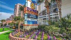 Letrero de Dunes Village Resort con un aviso de disponibilidad, un Captun's Cafe & Bar, y un jardín ajardinado con palmeras.