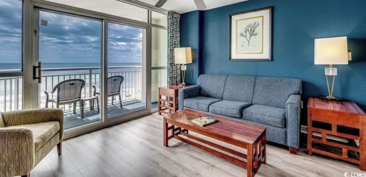 A room with a sofa, a coffee table, and a chair, a balcony with chairs, and a framed picture on the wall.