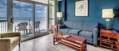 A room with a sofa, a coffee table, and a chair, a balcony with chairs, and a framed picture on the wall.