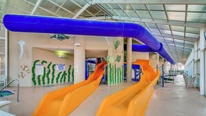 Parque acuático interior con dos toboganes, una piscina y un área cubierta con techo alto.