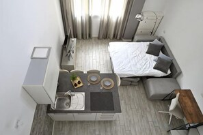 Ein kleines Hotelzimmer mit Küchenzeile, einem Bett mit grauer Bettwäsche, einem kleinen Esstisch und einem Fenster mit Vorhängen.