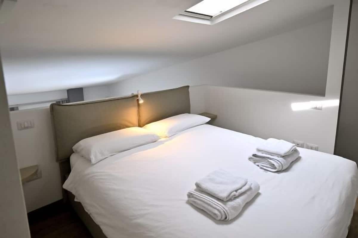 Una cama individual con sábanas blancas y dos toallas dobladas, un cabecero y un tragaluz arriba.