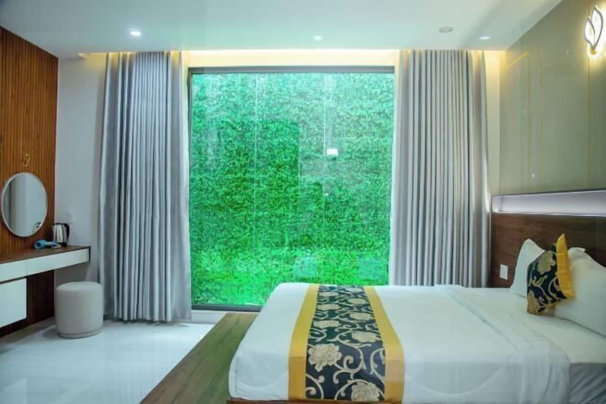 Une chambre d’hôtel moderne avec un grand lit, une coiffeuse en bois, un miroir rond et une vue sur un paysage verdoyant par la fenêtre.