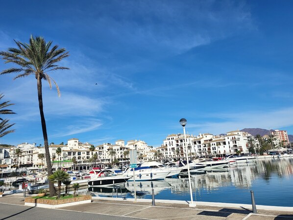 A marina with boats docked, surrounded by white buildings with balconies, palm trees, and a clear blue sky.