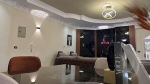 A modern hotel room with a glass table, a brown sofa, a bed with a tufted headboard, and a large window with a Netflix sign. - Chessboard Oasis - Elite 2BR apart by BC residence (New Cairo City)