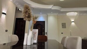 A modern dining area with a glass table, white chairs, and a vase with dried pampas grass. - Chessboard Oasis - Elite 2BR apart by BC residence (New Cairo City)