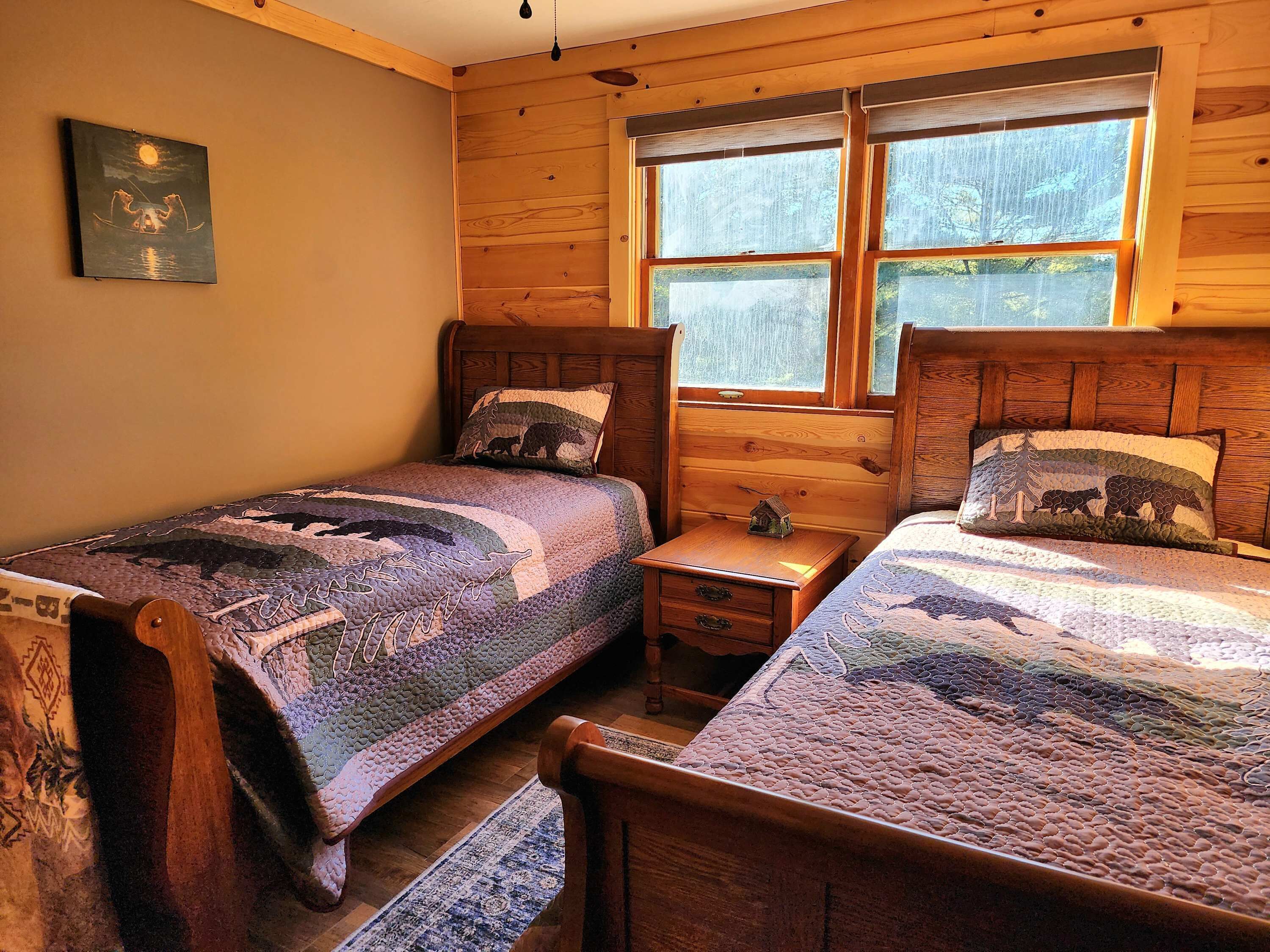 A cozy cabin room with two wooden beds, a small nightstand, and a window with a view of trees.