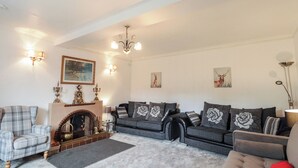 A living room with a fireplace, two sofas with floral cushions, a plaid armchair, and wall-mounted lamps.