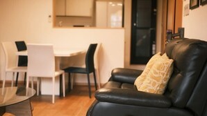 Un salon avec un canapé en cuir noir, une table basse en verre et un coin repas comprenant des chaises blanches et une table blanche.