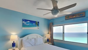 A bedroom with a bed, a ceiling fan, a window with a view of the ocean, and a painting of a beach scene.