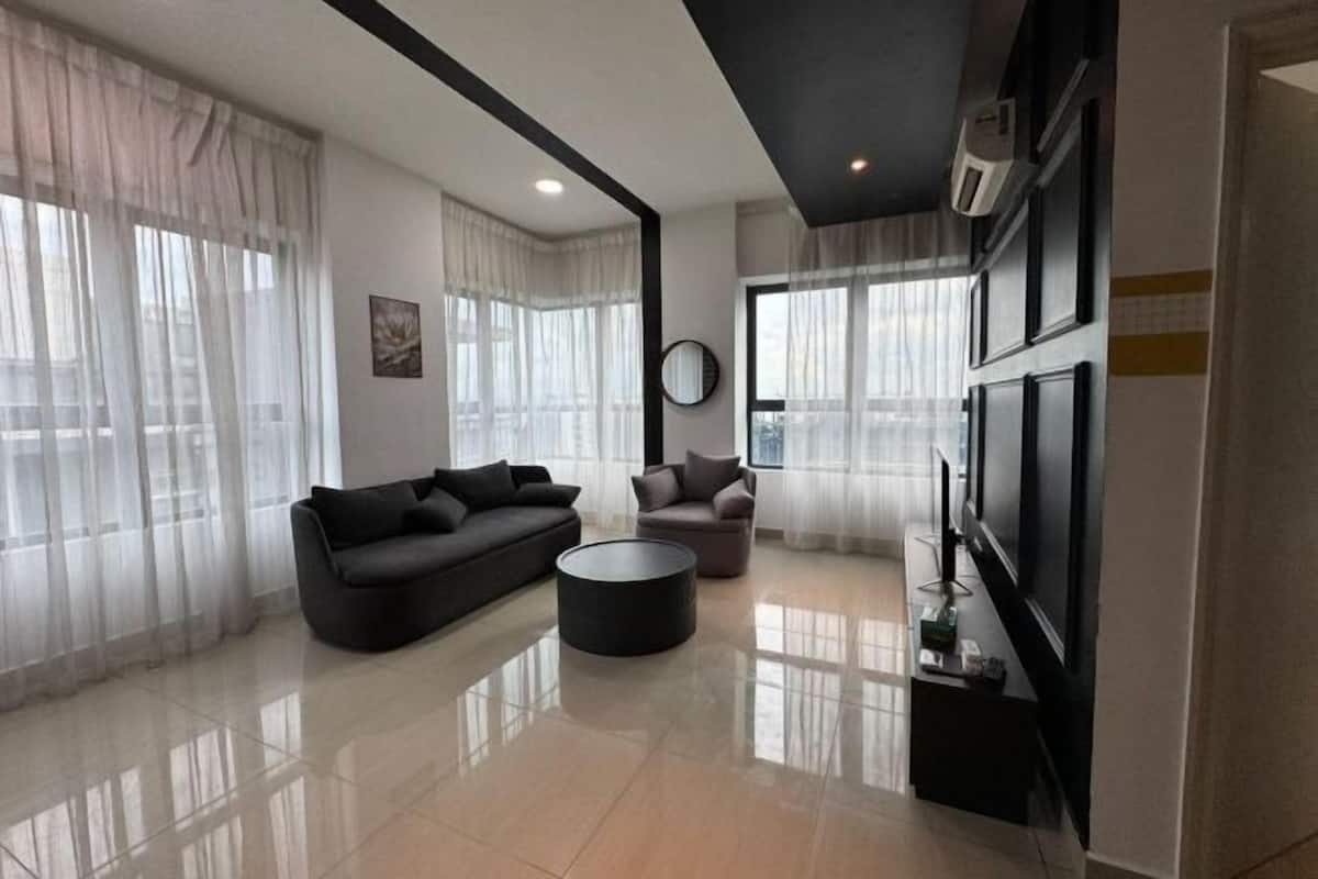 A modern living room with a sofa, armchair, and coffee table, large windows with sheer curtains, a flat-screen TV, and a wall-mounted air conditioning unit.