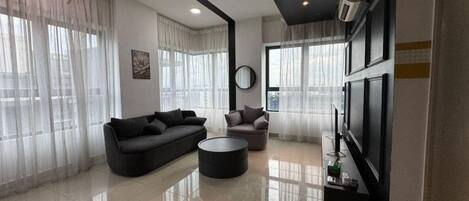 A modern living room with a sofa, armchair, and coffee table, large windows with sheer curtains, a flat-screen TV, and a wall-mounted air conditioning unit.