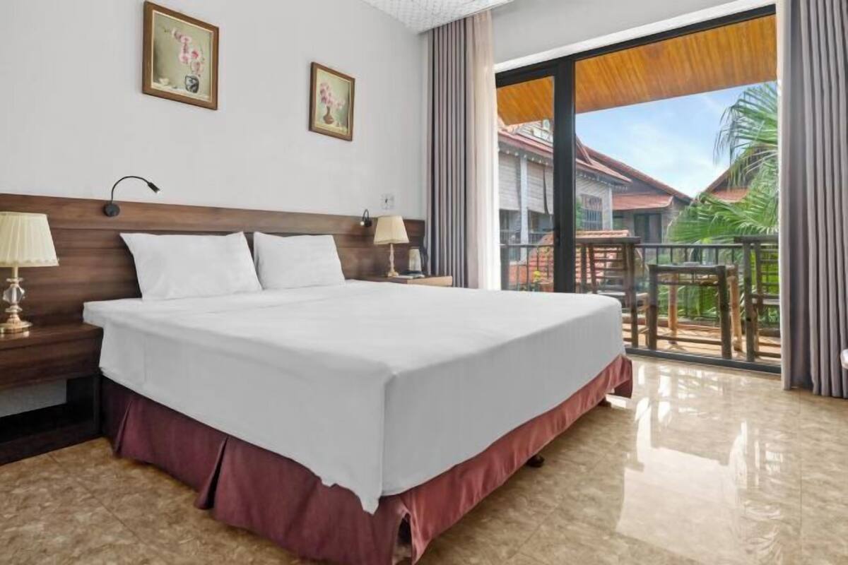 A hotel room with a large bed, a nightstand, a lamp, and a view of a balcony with outdoor furniture.