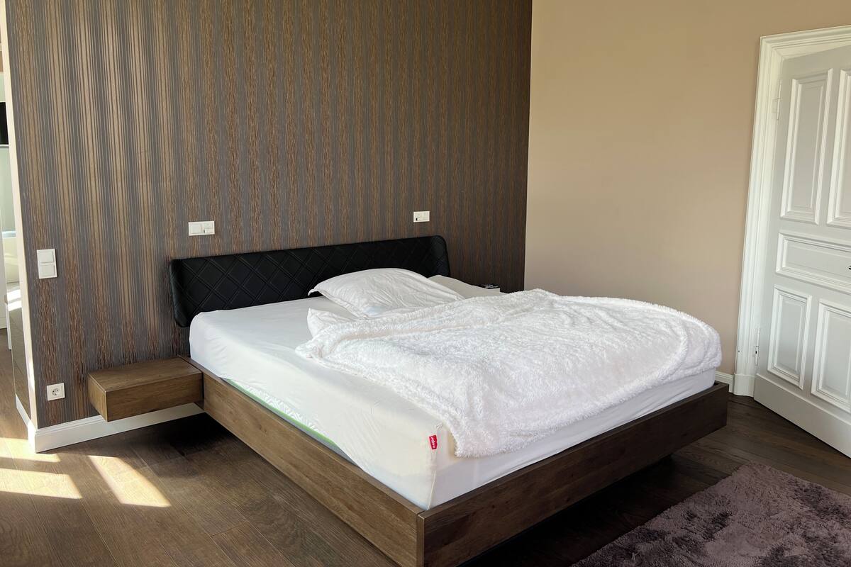 A neatly made bed with a white comforter and pillows, a wooden headboard, and a dark brown wall with vertical lines.