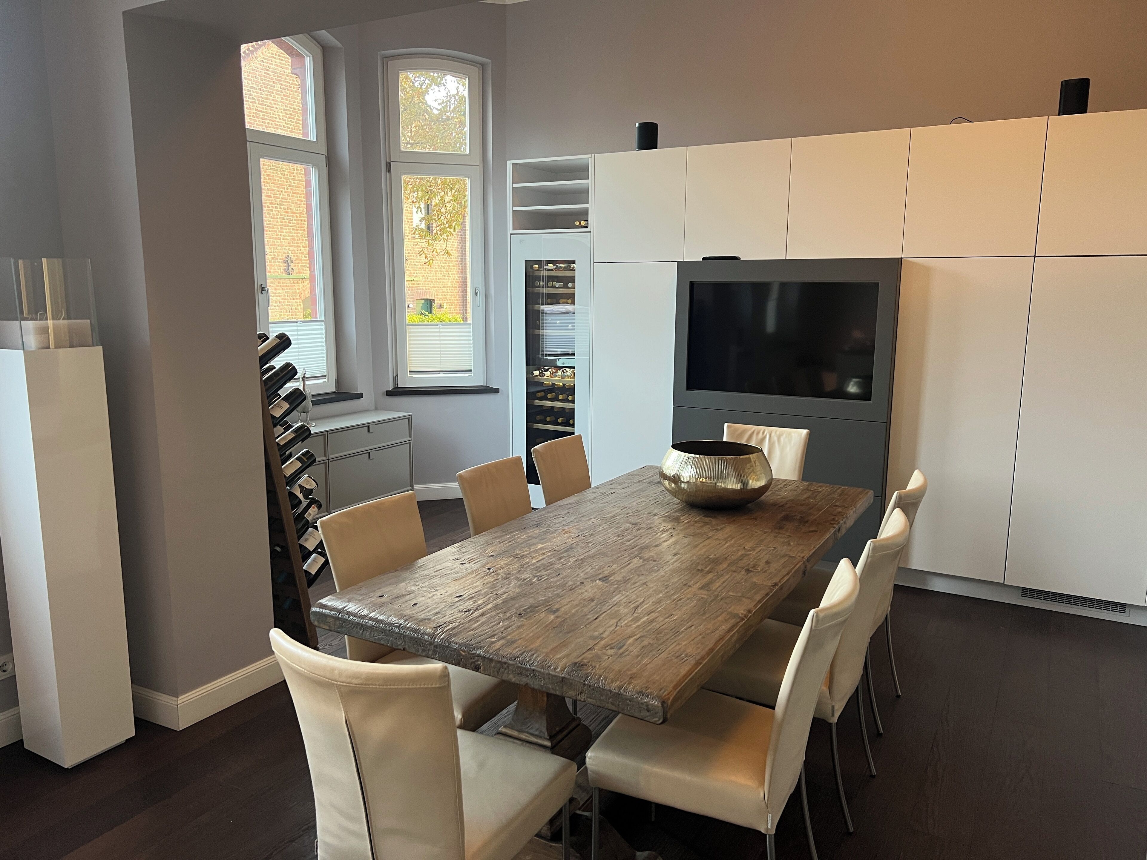 A modern kitchen with a wooden table, white chairs, a large TV, and a built-in oven.