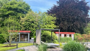 Ein moderner Außenbereich mit Holzterrasse, überdachter Sitzgruppe und einer Glasstruktur mit rotem Dach.